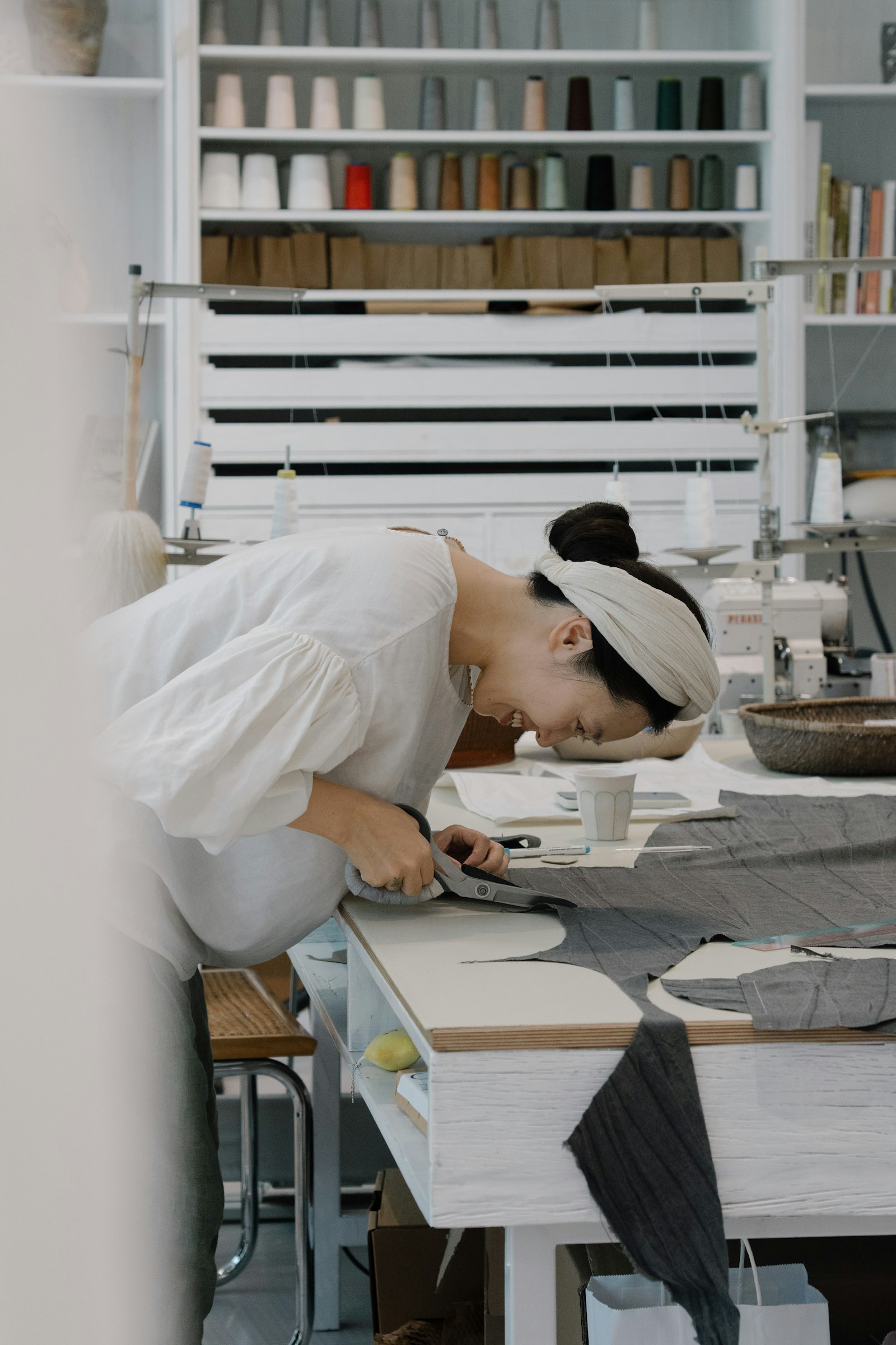 A maker in a bright atelier leaning over a cutting table with a sewing machine and thread cones behind