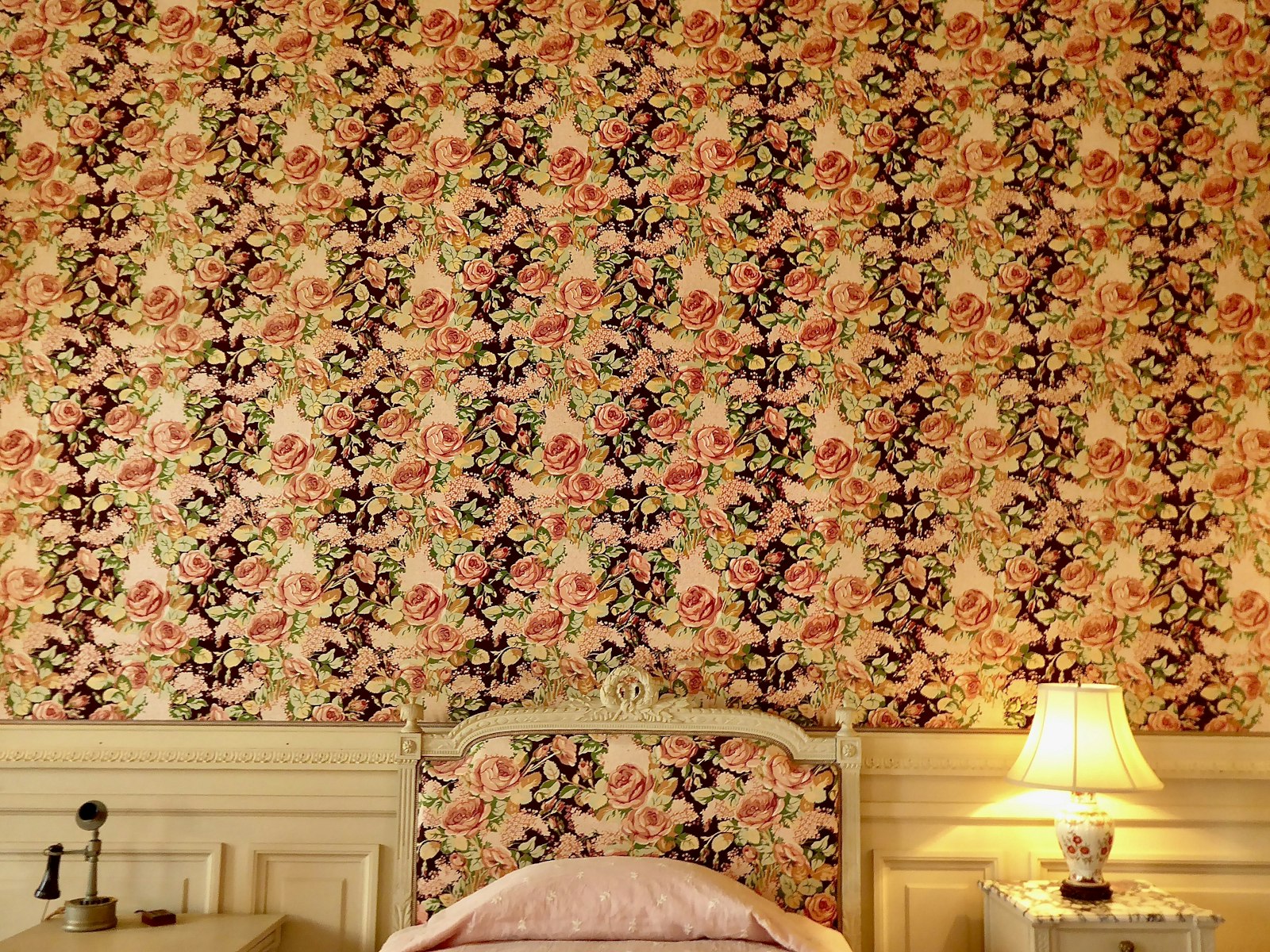 Peach and brown floral wallpaper behind a cream carved-wood headboard and brass lamp